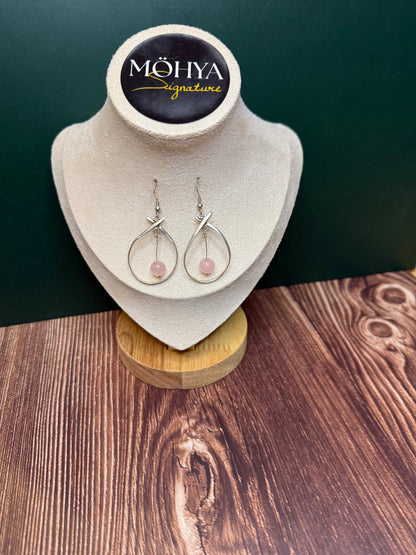 Par de brincos de prata com pedra rosa, design artesanal, expostos em suporte de veludo bege.