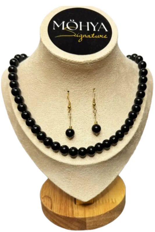 Conjunto de colar e brincos de contas pretas, exposto em suporte bege com base de madeira.